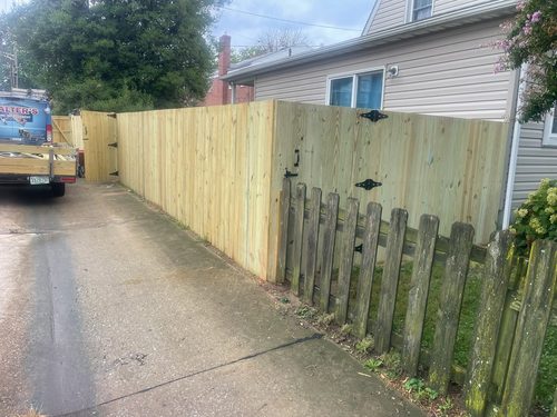 A new pressure-treated board fence and gate running along a driveway, beside the weathered picket fence it replaced